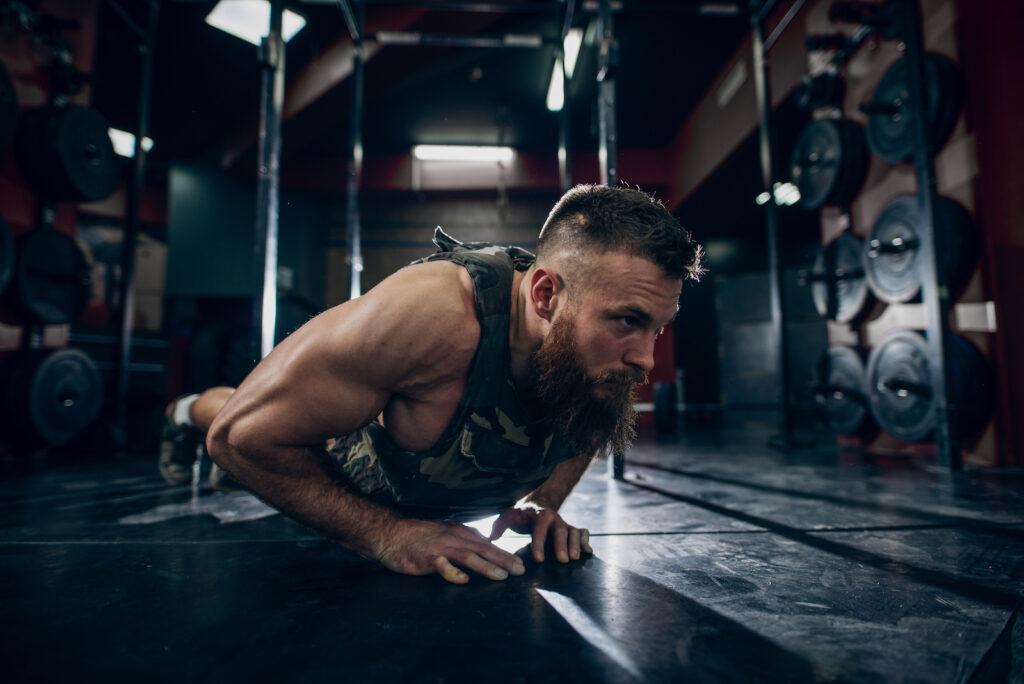Bearded man performing plank exercise on dark gym floor with weights visible in background