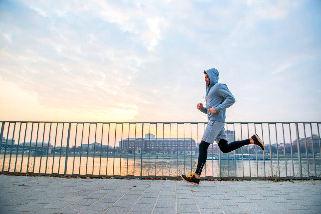 Person jogging on paved waterfront promenade with metal railing, urban skyline and water visible in background at sunrise or sunset