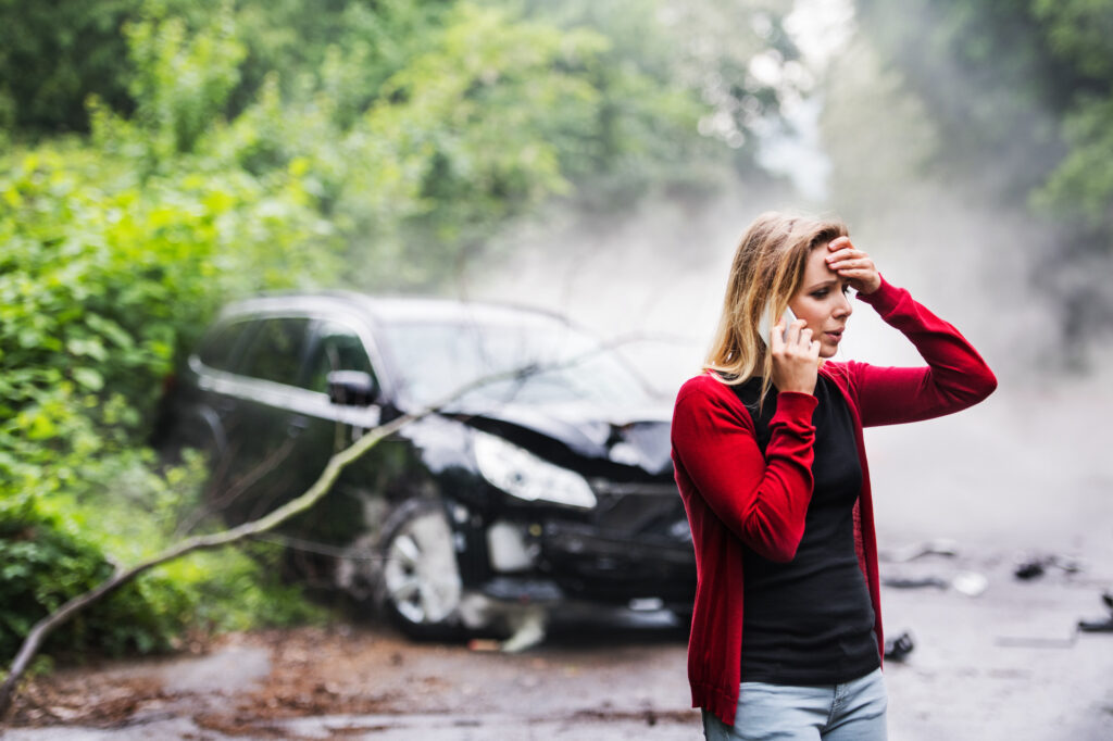 Woman in red cardigan on phone with damaged black car in background after accident