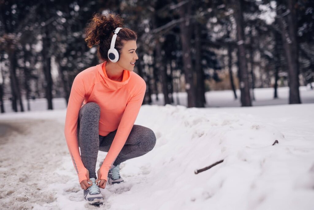 Woman in coral long-sleeve shirt and gray leggings stretches in snowy forest while wearing white headphones