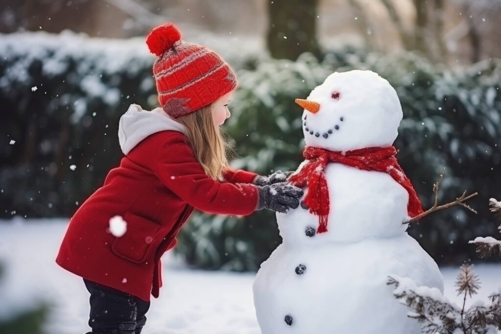 Child in red coat and knit hat touching a snowman wearing a red scarf during snowfall
