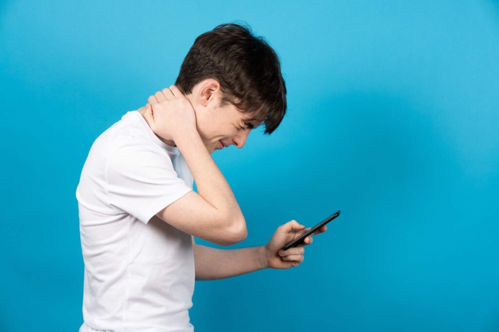 Teenage boy in white shirt holding smartphone at chest level, touching his neck in discomfort against a blue background.
