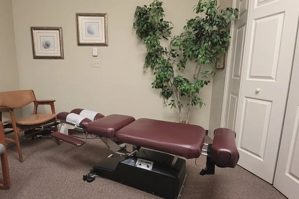 A quiet treatment room with a burgundy adjusting table, a potted tree and framed botanical prints