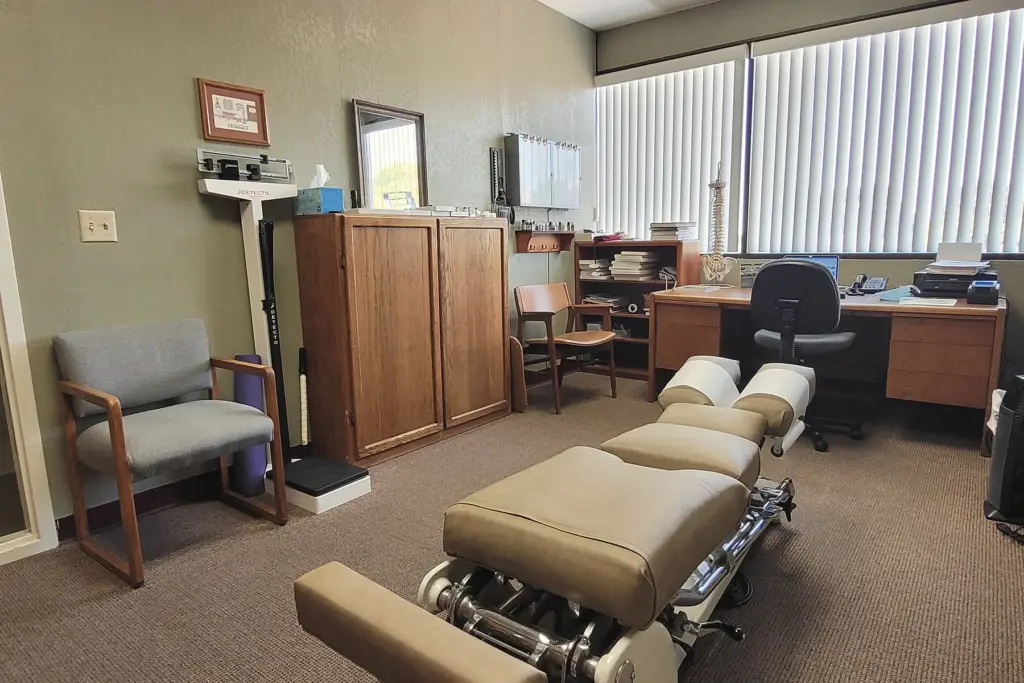 A bright treatment room with a padded adjusting table, a scale and a desk under tall windows